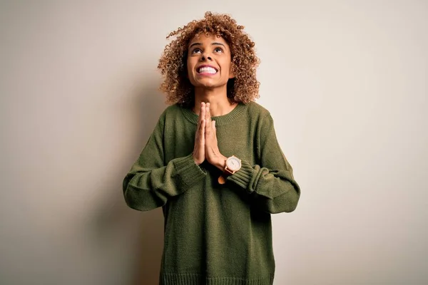 Beautiful african american woman with curly hair wearing casual sweater over white background begging and praying with hands together with hope expression on face very emotional and worried. Begging.