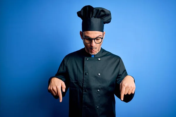 Young handsome chef man wearing cooker uniform and hat over isolated blue background Pointing down with fingers showing advertisement, surprised face and open mouth
