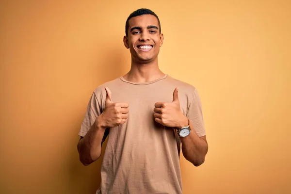 年轻英俊的非洲裔美国男人穿着休闲装站在黄色背景的成功标志上 用手做着积极的手势 微笑着竖起大拇指 快乐的样子 快乐的表情和胜利的姿态 — 图库照片