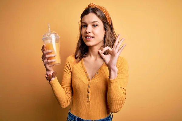年轻的金发姑娘喝着一杯清澈健康的橙汁 背景是黄色的 背景是孤立的 手握着手签 象征着完美 — 图库照片