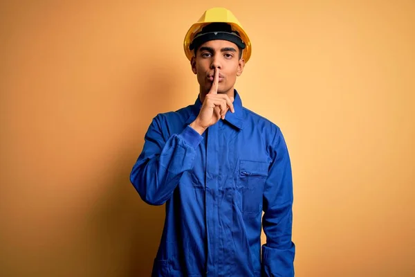 青い制服とセキュリティヘルメットを身に着けている若いハンサムなアフリカ系アメリカ人労働者の男性は唇に指で静かになるように求めている 沈黙と秘密の概念 — ストック写真