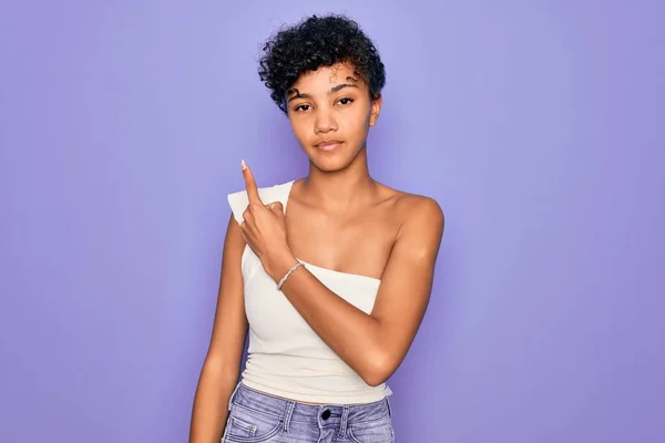 年轻美丽的非洲裔美国女人 穿着休闲的T恤 背景是紫色的 手指头指向广告的侧面 严肃而沉着 — 图库照片