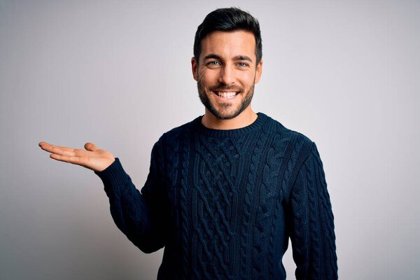 Young handsome man with beard wearing casual sweater standing over white background smiling cheerful presenting and pointing with palm of hand looking at the camera.