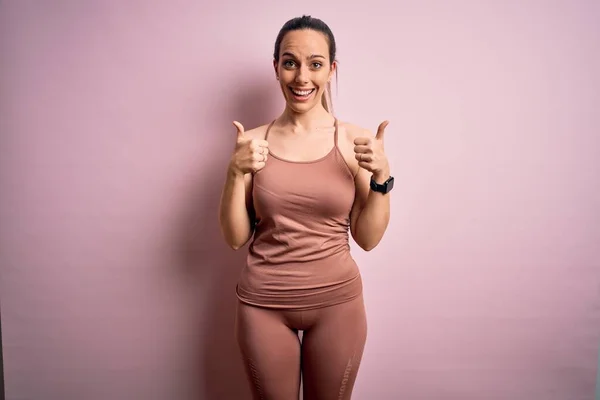 Young blonde fitness woman wearing sport workout clothes over pink isolated background success sign doing positive gesture with hand, thumbs up smiling and happy. Cheerful expression and winner gesture.