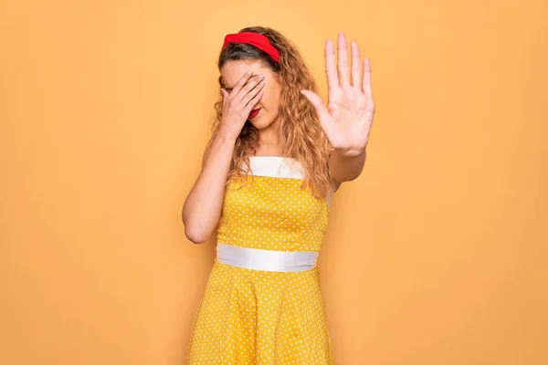 Beautiful blonde pin-up woman with blue eyes wearing diadem standing over yellow background covering eyes with hands and doing stop gesture with sad and fear expression. Embarrassed and negative concept.