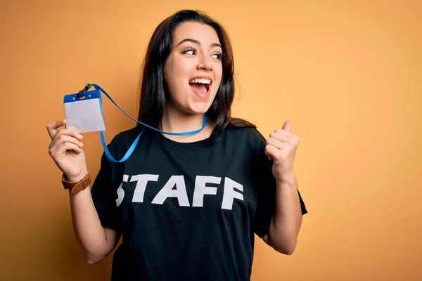 Young brunette worker woman wearing staff t-shirt as uniform showing id card pointing and showing with thumb up to the side with happy face smiling