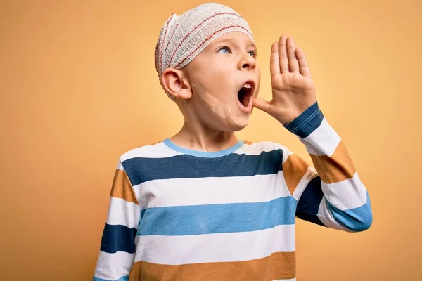 Young little caucasian kid injured wearing medical bandage on head over yellow background shouting and screaming loud to side with hand on mouth. Communication concept.