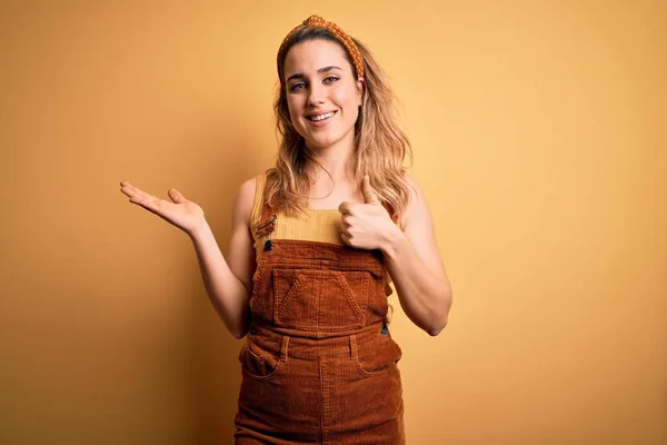 Young beautiful blonde woman wearing overalls and diadem standing over yellow background Showing palm hand and doing ok gesture with thumbs up, smiling happy and cheerful