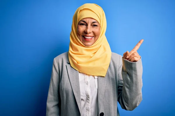 中年黑发女商人头戴穆斯林式的传统头巾 头戴蓝色背景 脸上挂着大大的笑容 手指头指向旁边看相机 — 图库照片