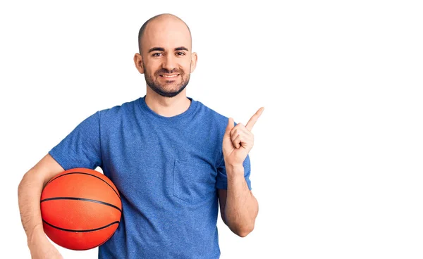 年轻英俊的男子拿着篮球开心地笑着 手指头指向旁边 — 图库照片
