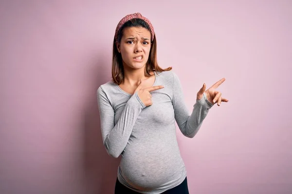 若いです美しいですティーン女の子妊娠期待赤ちゃん上の隔離されたピンクの背景ポインティング脇心配と神経で両方の手 懸念と驚きの表現 — ストック写真