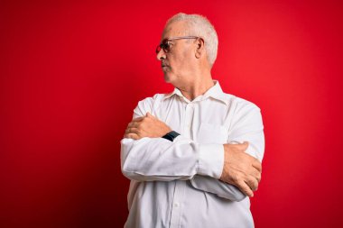Orta yaşlı yakışıklı, beyaz gömlekli, kırmızı arka planda gözlüklü, kolları çapraz ve kendinden emin bir adam.