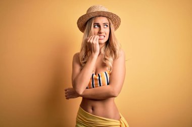 Bikini giyen, sarı arka planda şapka takan genç ve güzel sarışın kadın elleri ağız yiyen tırnaklarla stresli ve gergin görünüyor. Endişe sorunu.