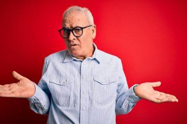 Orta yaşlı, yakışıklı, beyaz bir adam kırmızı arka planda sıradan çizgili gömlek ve gözlük takıyor. Hiçbir şeyden habersiz ve kolları açık, hiçbir fikri yok..