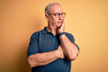 Orta yaşlı yakışıklı beyaz adam sarı arka plan üzerine günlük polo ve gözlük takıyor. Kollarını kavuşturup depresyondan sıkılmış ve yorgun görünüyor..
