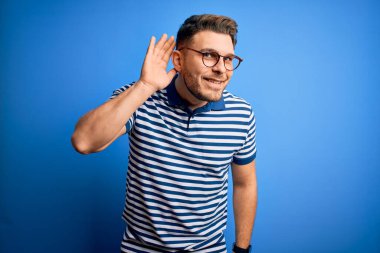 Mavi gözlü, gözlüklü, mavi arkaplanlı, sıradan çizgili tişörtlü, ağzı kulaklarında, dedikoduları ve dedikoduları dinleyen genç bir adam. Sağırlık kavramı.