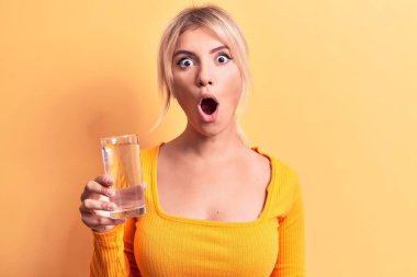 Sarışın, güzel bir kadın, izole edilmiş sarı arka planda bir bardak su içiyor. Şaşırmış ve şaşırmış bir şekilde ağzı açık, inançsız bir yüz.