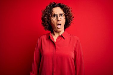 Orta yaşlı, kıvırcık saçlı, normal gömlekli, kırmızı arka planda gözlüklü, kuşkucu ve alaycı bakışlı, ağzı açık, şaşırmış bir kadın.