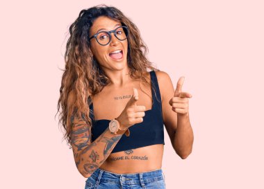 Sıradan kıyafetler ve gözlük takan, mutlu ve komik yüzlü, parmaklarını kameraya gösteren Latin kökenli genç bir kadın. iyi enerji ve titreşimler. 