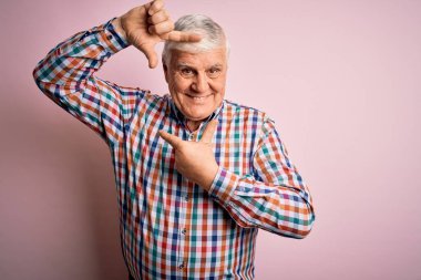 Üst düzey yakışıklı, parlak gömlekli, pembe arka planda izole edilmiş gülümseyen, mutlu yüzlü elleri ve parmakları olan bir çerçeve yapan bir adam. Yaratıcılık ve fotoğrafçılık kavramı.