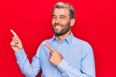 Genç, sarışın, gündelik gömlek giyen, izole edilmiş kırmızı arka planda duran, gülümseyen ve kameraya bakan, iki elini ve parmağını kenarı gösteren bir adam..