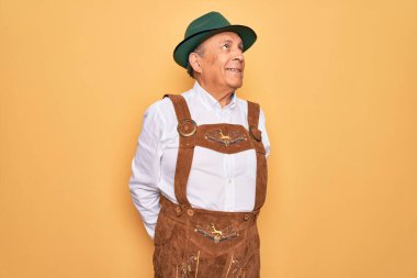 Kıdemli gri saçlı, Alman geleneksel Ekim Festivali kıyafeti giyen, sarı arka planda gülümseyen ve düşüncelere dalmış bir adam..