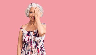 Kıdemli gri saçlı kadın, günlük kıyafetler giyiyor, bir gözünü eliyle kapatıyor, yüzünde kendine güvenen bir gülümseme ve şaşırtıcı bir duygu var.. 