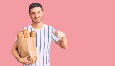 Elinde ayı olan yakışıklı genç adam elinde ekmek torbasıyla mutlu ve gururlu gülümseyen birini işaret ediyor. 