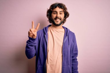 Sakallı, pembe arka planda sıradan bir kazak giyen genç yakışıklı sporcu kameraya bakıp zafer işareti yapıyor. İki numara..