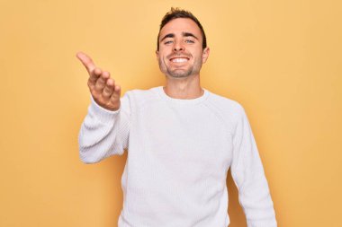 Mavi gözlü, sıradan bir süveter giyen, sarı arka planda duran yakışıklı genç bir adam dostça bir selamlama ve hoş geldin hediyesi olarak el sıkışmayı sunuyor. Başarılı bir iş..