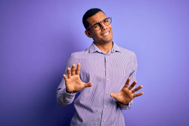 Yakışıklı Afro-Amerikan erkeği çizgili gömlek ve gözlük takıyor mor arka planda, tiksindirici ve tiksindirici yüz ifadesi takınıyor çünkü tiksindirici tepki veriyor..