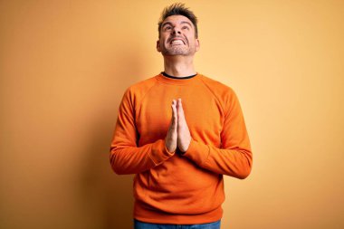 Turuncu kazak giyen genç yakışıklı adam izole edilmiş sarı arka planda duruyor. Elleri bağlı yalvarıyor ve dua ediyor. Yüzünde umut ifadesi var. Yalvarıyorum..