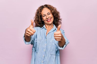 Orta yaşlı güzel bir kadın pembe arka plan üzerinde dikilmiş olumlu jestler yapıyor, gülümsüyor ve başarı için mutlu. Kazanan jest.