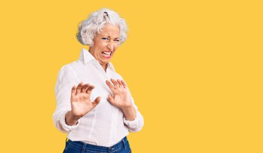 Kıdemli, gri saçlı, günlük kıyafetler giyen, tiksindirici ifadeler takınan, tiksindirici, tiksindirici bakışlar atan bir kadın. Eller havada. 