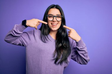 Mor izole arka planda gözlük takan genç esmer kadın neşeli gülüyor ve parmaklarıyla ve ağzıyla işaret ediyor. Diş sağlığı konsepti.
