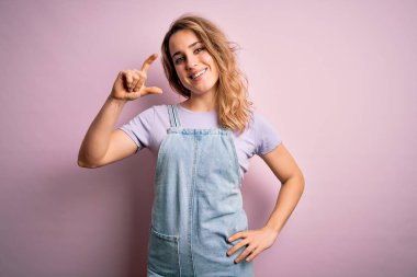 Genç, güzel, sarışın bir kadın pembe arka planda dikilmiş rahat kot pantolon giyiyor gülümsüyor ve el hareketleriyle küçük beden işareti yapıyor parmakları bakıyor ve kameraya bakıyor. Ölçü kavramı.