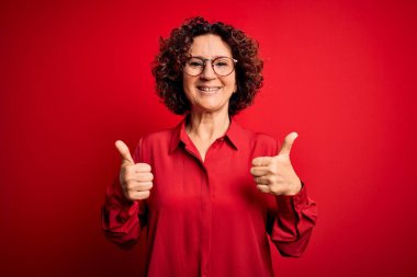 Orta yaşlı güzel kıvırcık saçlı kadın kırmızı arka plan işareti üzerine günlük gömlek ve gözlük takıyor. Elleriyle olumlu jest yapıyor. Gülümsüyor ve mutlu. Neşeli ifade ve kazanan jest.