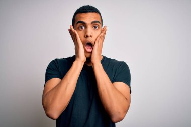 Beyaz arka planda duran sıradan tişörtler giyen genç ve yakışıklı Afro-Amerikan adam korkmuş ve şok olmuş, şaşırmış ve şaşırmış bir halde.