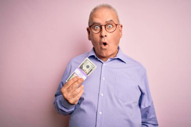 Orta yaşlı, ağarmış bir adam pembe arka planda bir deste banknot tutuyor. Şaşırmış bir yüz ifadesinden korkuyor ve heyecanlanıyor.