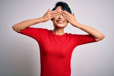 Genç, güzel Asyalı bir kız, günlük kırmızı tişört giyip izole edilmiş beyaz arka planda duruyor. Gözleri kaplıyor, neşeli ve komik gülümsüyor. Kör kavram.