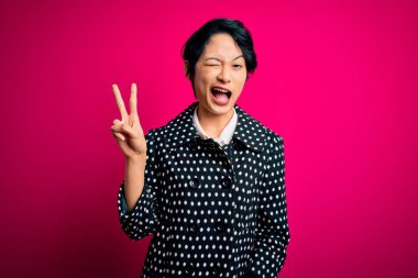 Genç ve güzel Asyalı bir kız, günlük ceket giyip pembe arka planda dikiliyor. Gülümsüyor, kameraya göz kırpıyor ve parmaklarıyla zafer işareti yapıyor. İki numara..