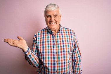 Üst düzey yakışıklı, parlak gömlekli, soyutlanmış pembe arka planda neşeli bir şekilde gülümseyen ve avuç içi kameraya bakan bir adam..