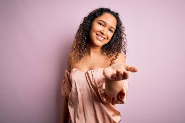 Kıvırcık saçlı, pembe arka planda dikilen günlük tişörtlü neşeli gülümsemeli, yardım eli uzatan ve kabul gören genç bir kadın..