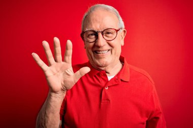 Gri saçlı, gözlüklü, kırmızı arka planda günlük tişörtlü, kendinden emin ve mutlu bir şekilde gülümseyen beş numarayı işaret eden son sınıf öğrencisi..