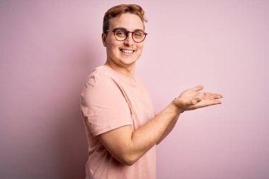 Genç, kızıl saçlı, sıradan bir tişört giyen, pembe arka planda duran, avuç içi açık, fotokopi alanını gösteren, mutlu bir şekilde gülümseyen bir reklam sunan genç bir adam.
