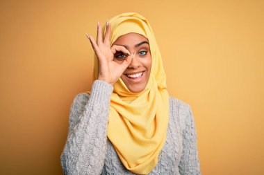 Genç ve güzel Afro-Amerikalı kız izole edilmiş sarı arka plan üzerine Müslüman başörtüsü takıyor. El hareketleriyle gülümsüyor, gözleriyle mutlu yüzle bakıyor..