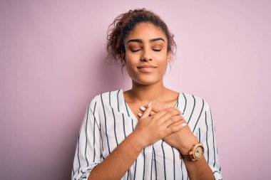 Genç ve güzel Afro-Amerikalı kız pembe arka planda duran çizgili tişört giyiyor elleri göğsünde, gözleri kapalı ve yüzünde minnet dolu bir jest var. Sağlık kavramı.