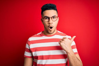 Sıradan çizgili tişört ve gözlük takan genç yakışıklı adam izole edilmiş kırmızı arka planda el ile işaret ederek şaşırdı, ağzı açık hayret içinde kaldı..
