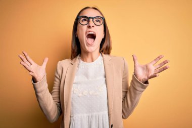 Ceket ve gözlük takan genç ve güzel iş kadını sarı arka planda çılgınca bağırıyor ve agresif bir ifade ve kollarla bağırıyor. Hayal kırıklığı konsepti.