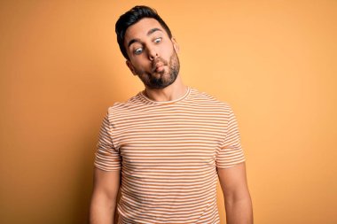 Sakallı, sarı arka planda günlük çizgili tişört giyen genç yakışıklı adam dudaklı balık suratı yapıyor, çılgınca ve komik bir jest. Komik ifade.